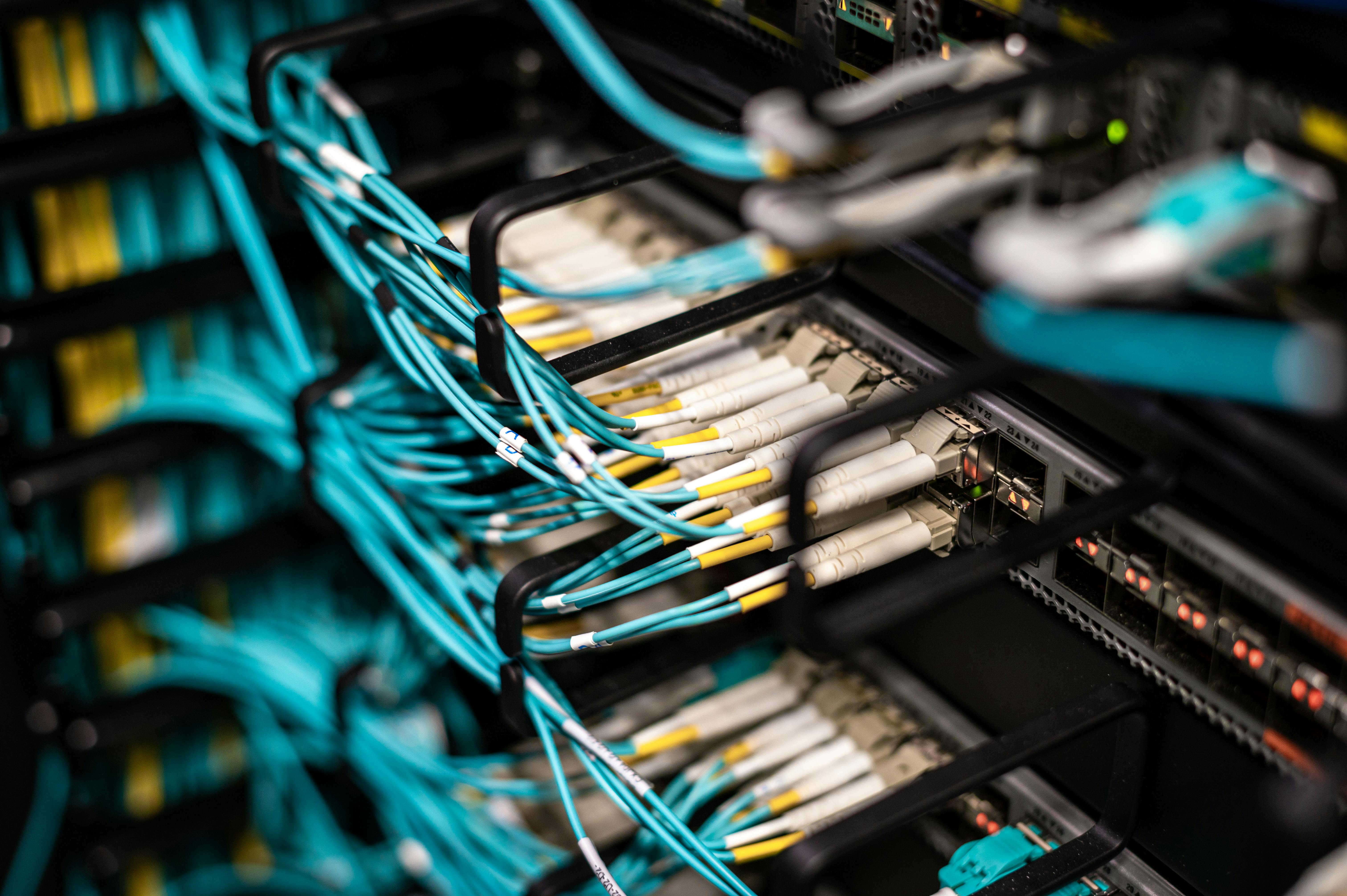 Close-up of a network rack with multiple rows of aqua-colored fiber optic cables connected to white and yellow connectors, neatly routed through black cable management arms in a well-maintained data center or server room