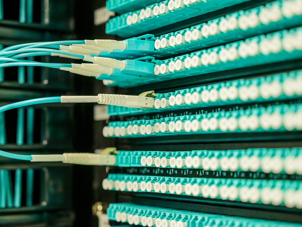 Close-up of a network rack with multiple rows of aqua-colored fiber optic cables connected to white and blue connectors, used for high-speed data transfer in a data center or server room.