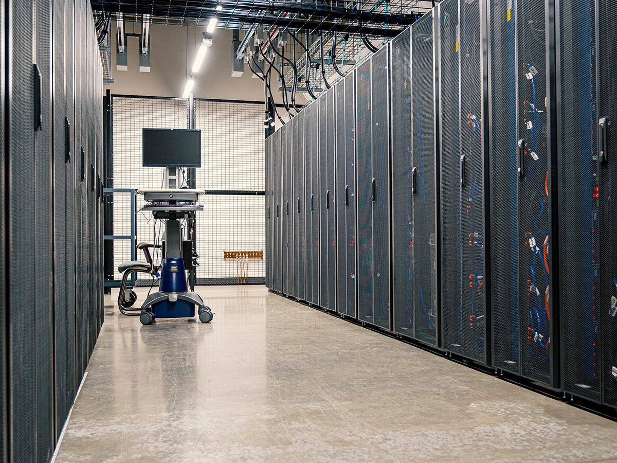 A data center aisle with rows of server racks on both sides, featuring networking equipment and cables. A mobile workstation with a monitor, keyboard, and nearby chair is positioned in the center. Overhead cable trays and lighting fixtures are visible on the ceiling