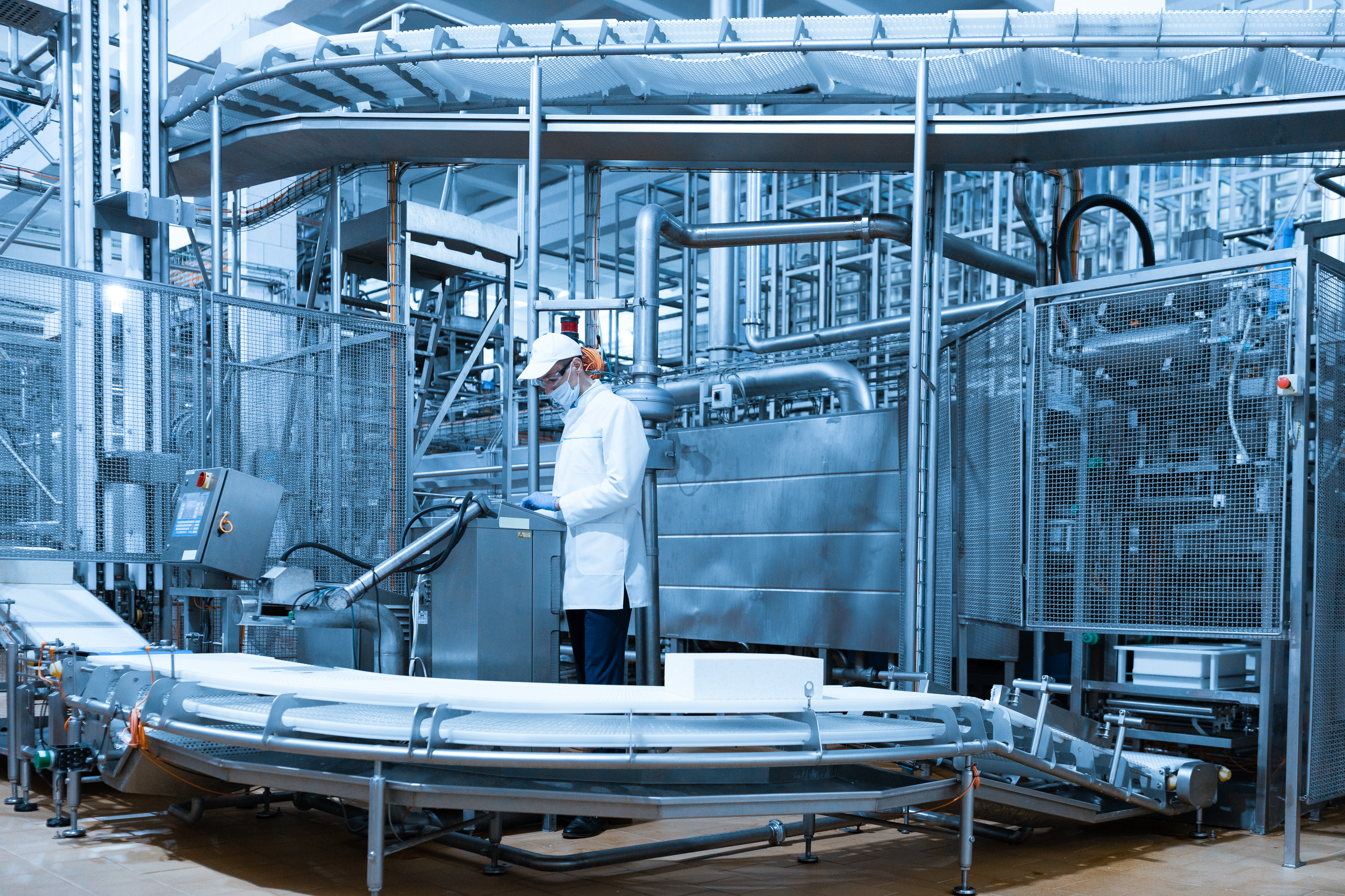 Industrial environment with a worker in a white lab coat and cap operating machinery on an automated conveyor belt system, surrounded by metal structures, pipes, and equipment.