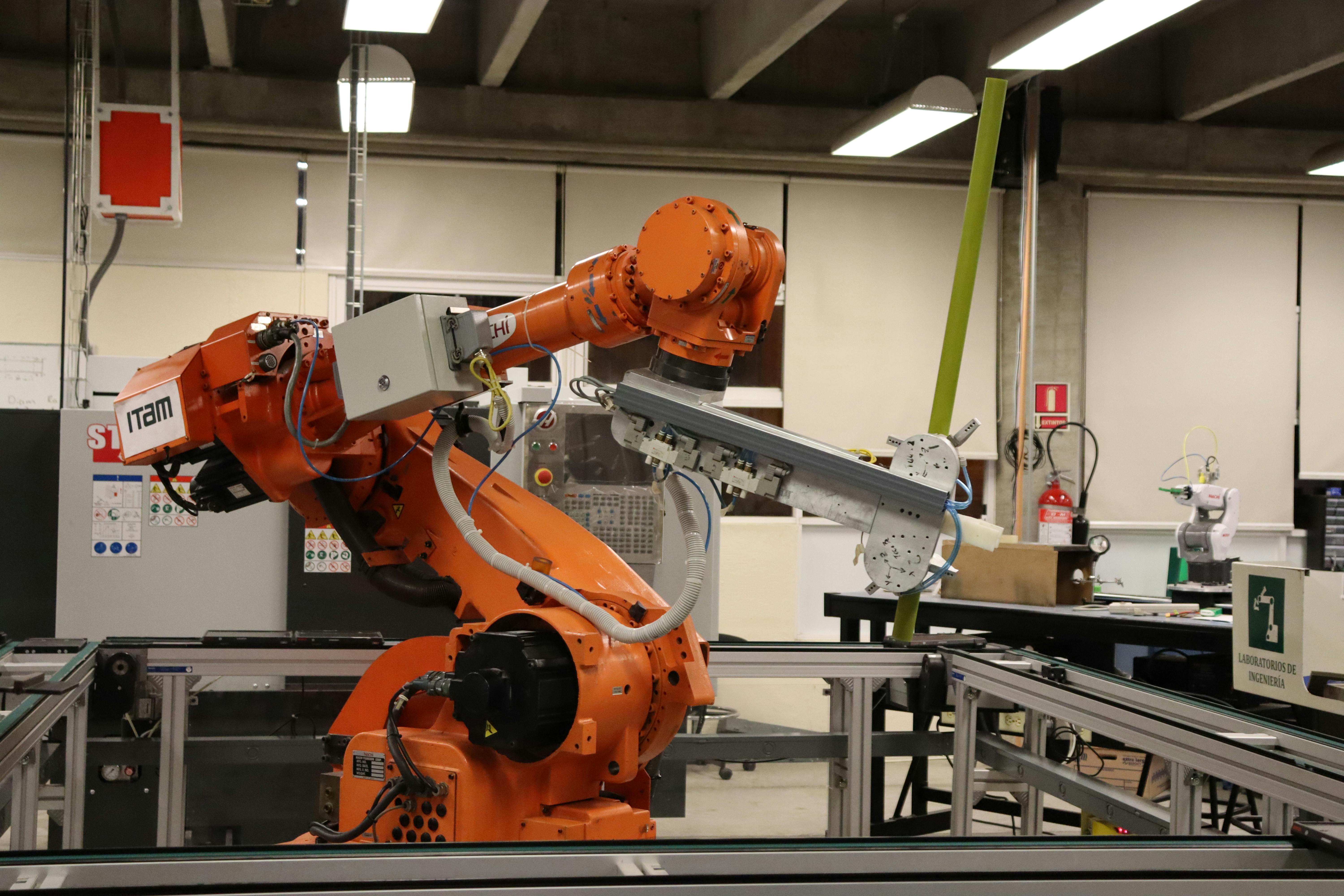 Industrial setting with an orange robotic arm labeled 'IRAM' operating on an automated assembly line, surrounded by machinery and equipment under bright overhead lighting.