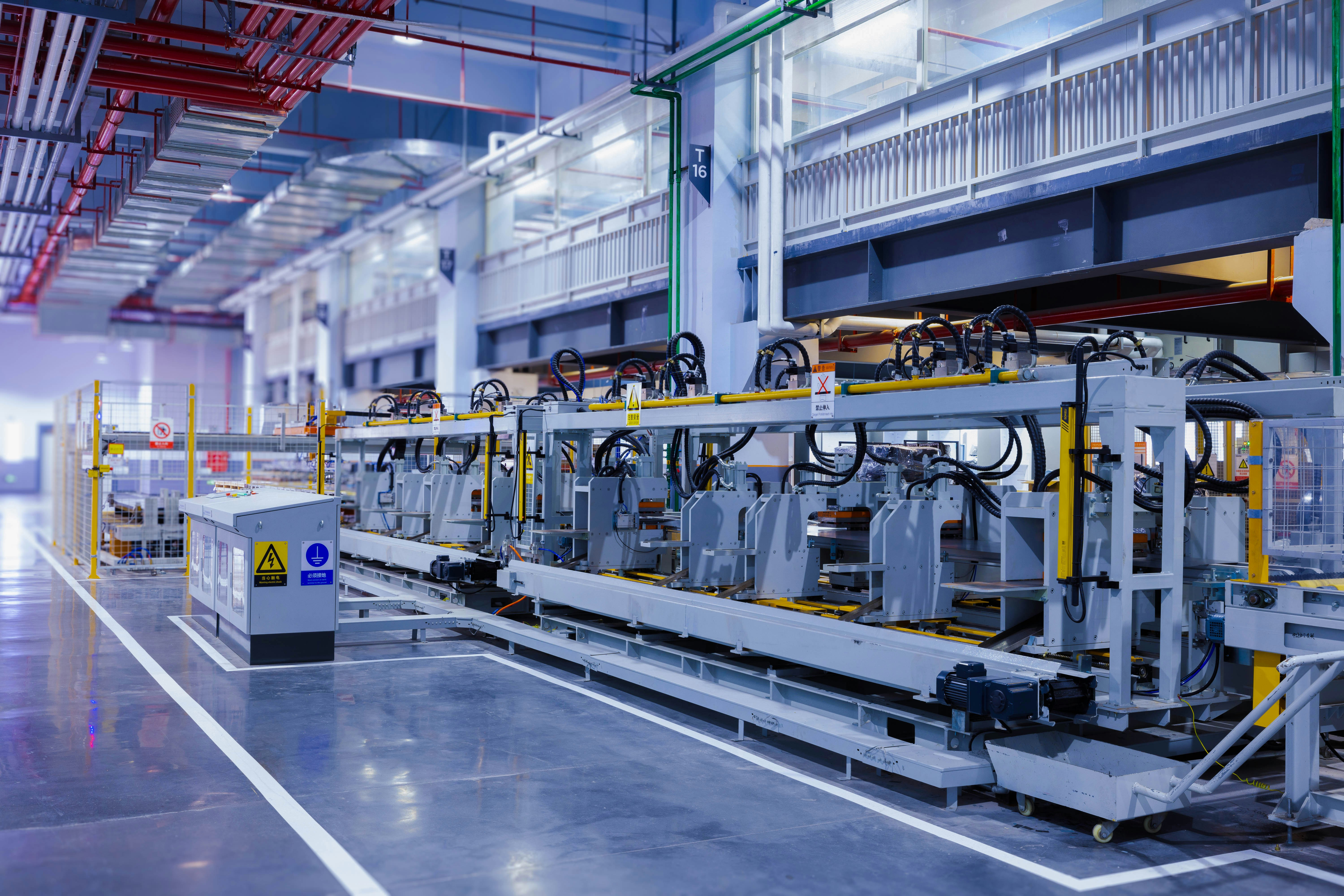 Industrial factory with a long automated assembly line featuring robotic arms and machinery, enclosed by safety barriers and warning signs, under bright overhead lighting and visible ceiling piping.