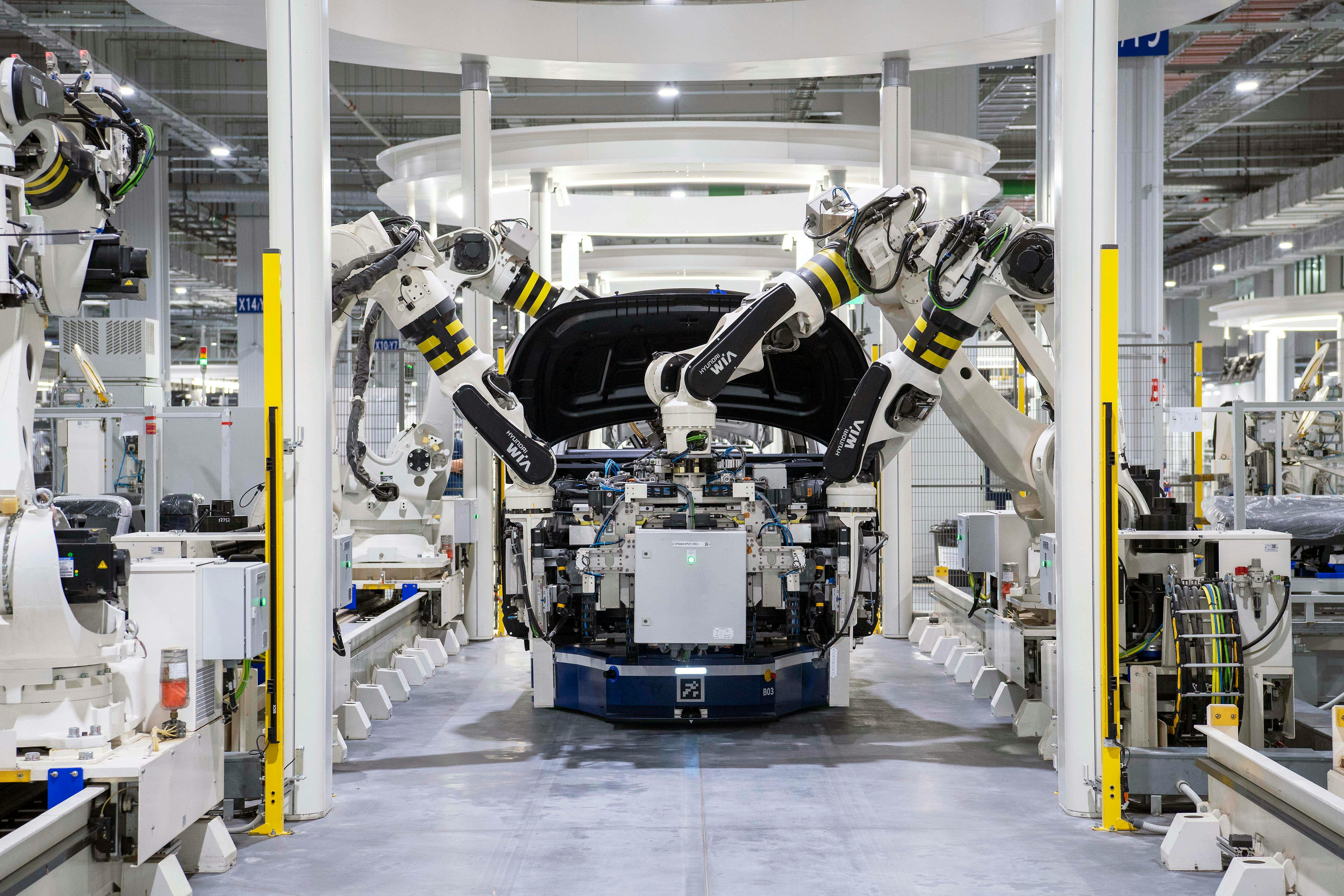Automated factory assembly line with robotic arms working on a car chassis, surrounded by industrial equipment and illuminated by bright overhead lighting.