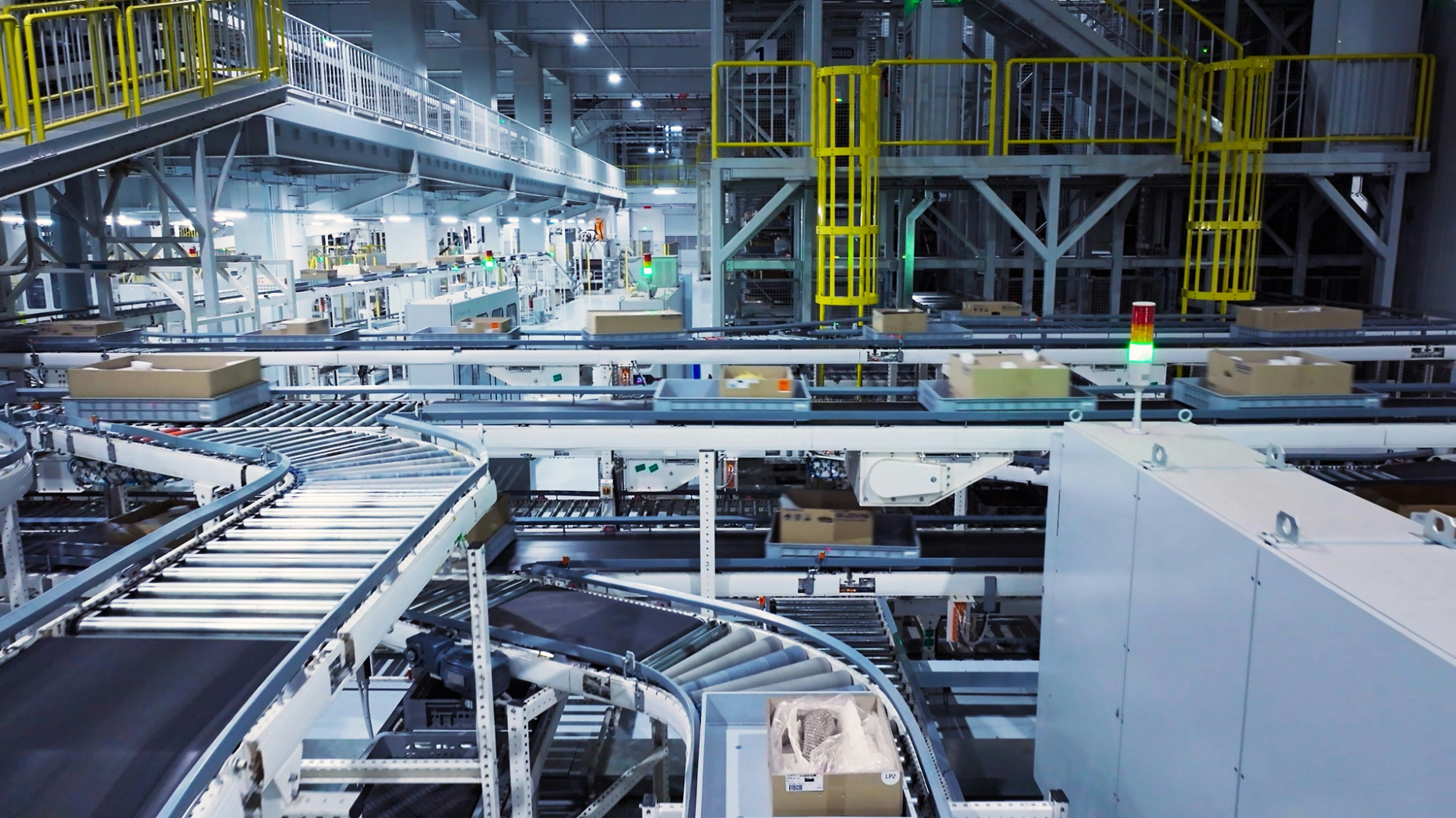 Large industrial warehouse with an automated conveyor belt system moving packages, surrounded by metal structures, safety railings, and bright overhead lighting.