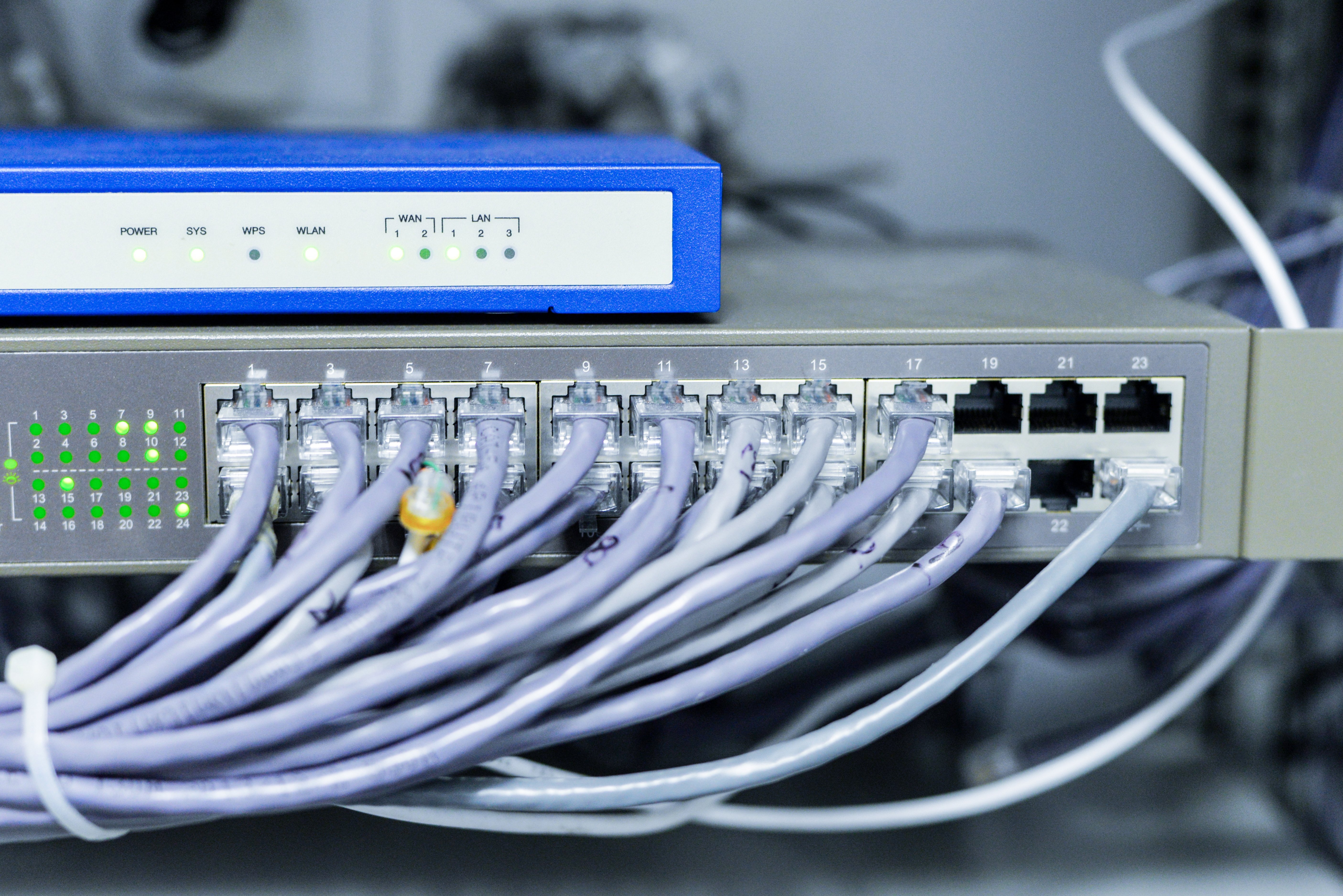 Close-up of a network switch with Ethernet cables connected to ports numbered 1 to 24, and a blue device above it displaying indicator lights labeled POWER, SYS, WPS, WLAN, and LAN.