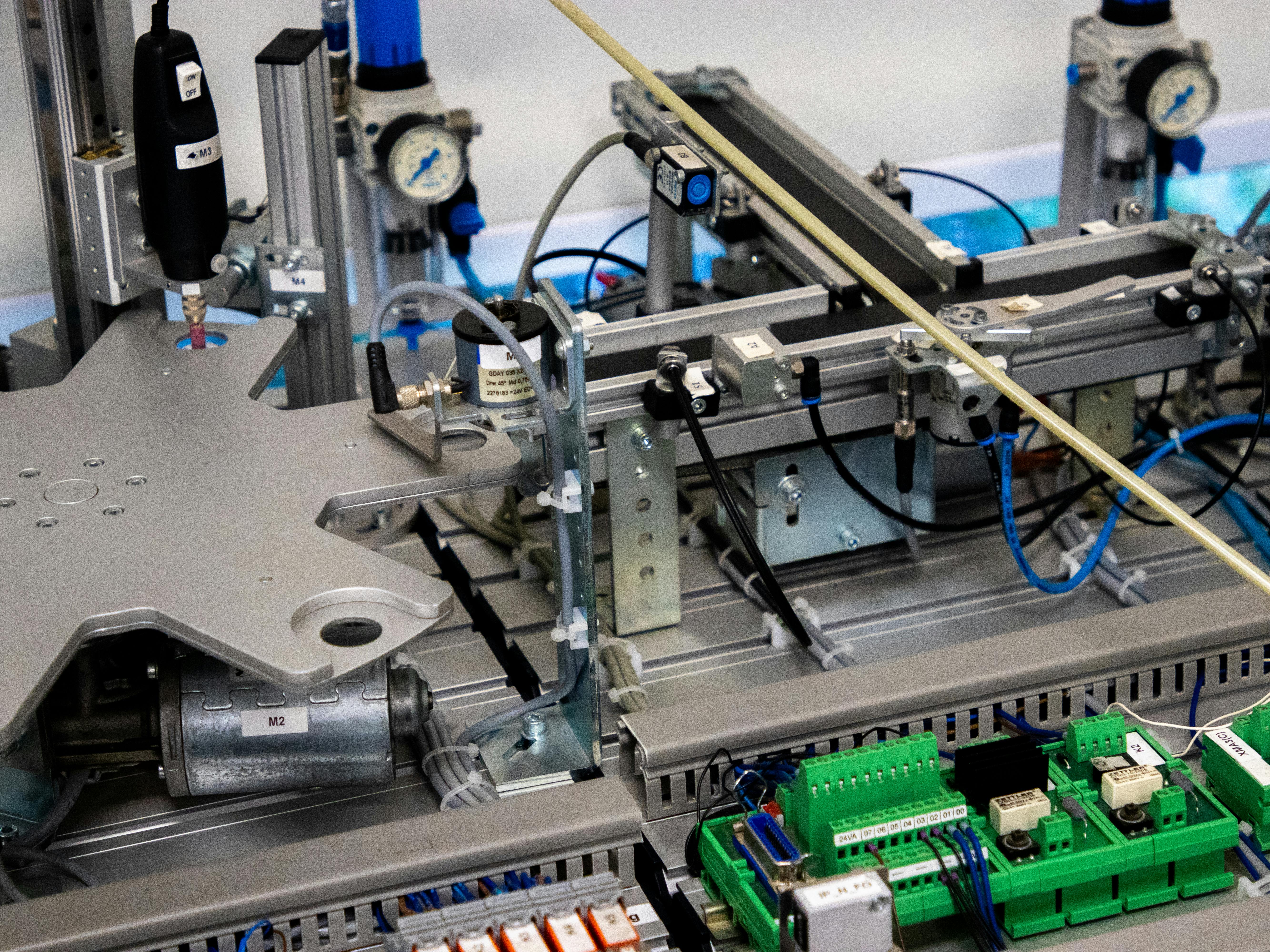 Close-up of an industrial automation system with metal frames, pneumatic cylinders, pressure gauges, green circuit boards, and connected wiring and sensors.